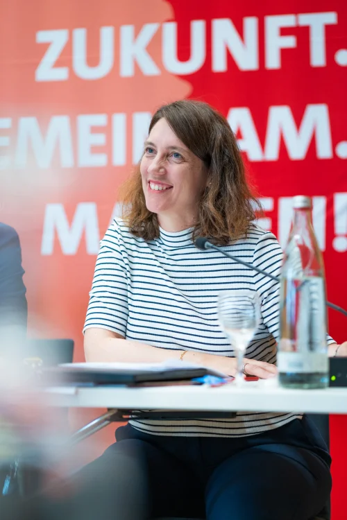 Dorothea Kliche-Behnke sitzt lächelnd vor einem rotem SPD‑Banner, vor ihr liegen Mikrofon, Unterlagen und ein Wasserglas auf dem Tisch.
