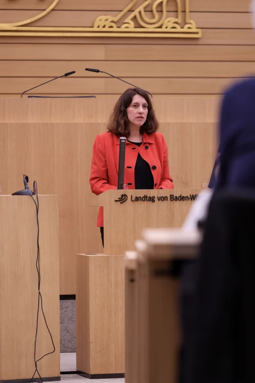 Dorothea Kliche-Behnke spricht im roten Blazer spricht Rednerpult im Plenarsaal des Landtags von Baden‑Württemberg, vor holzvertäfelter Wand mit Landeswappen.